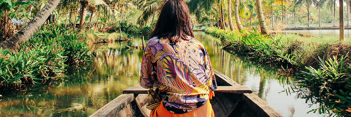 Eine Frau mit buntgemusterter Bluse sitzt in einem hölzernen Kanu und ist von hinten zu sehen. Sie fährt über einen mit Palmen gesäumten Fluss.