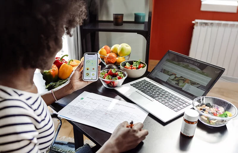 Eine Frau sitzt mit einem Smartphone am Tisch. Vor ihr liegen ein Ernährungsplan und gesunde Lebensmittel.