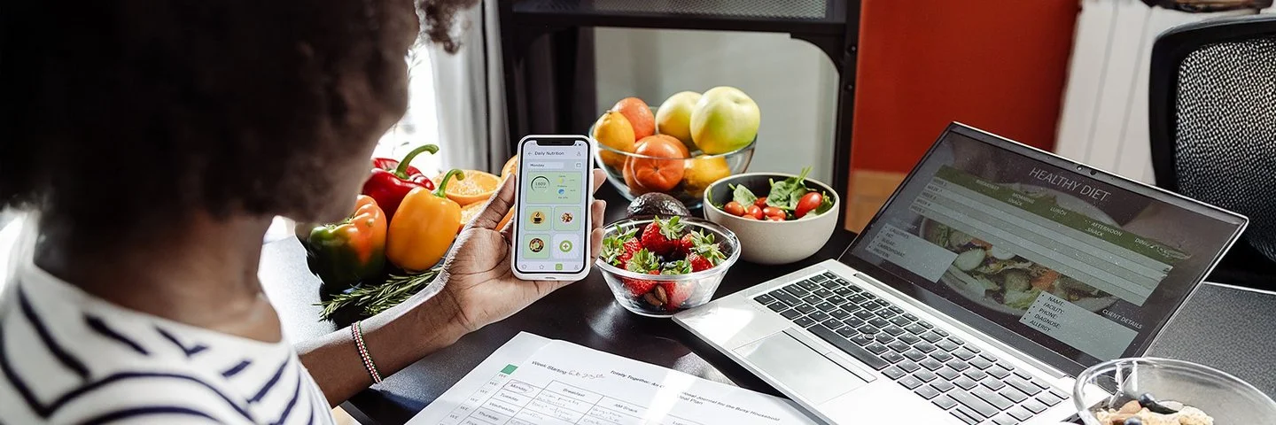 Eine Frau sitzt mit einem Smartphone am Tisch. Vor ihr liegen ein Ernährungsplan und gesunde Lebensmittel.