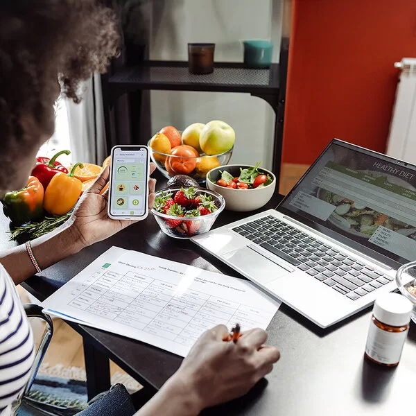 Eine Frau sitzt mit einem Smartphone am Tisch. Vor ihr liegen ein Ernährungsplan und gesunde Lebensmittel.