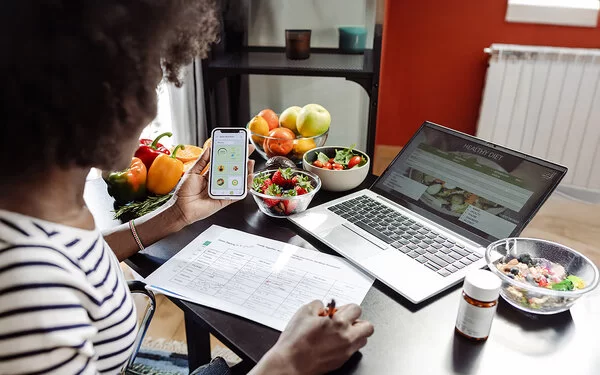 Eine Frau sitzt mit einem Smartphone am Tisch. Vor ihr liegen ein Ernährungsplan und gesunde Lebensmittel.