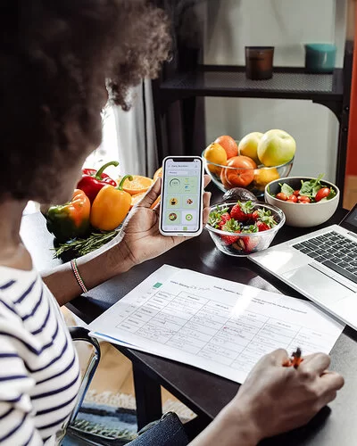 Eine Frau sitzt mit einem Smartphone am Tisch. Vor ihr liegen ein Ernährungsplan und gesunde Lebensmittel.