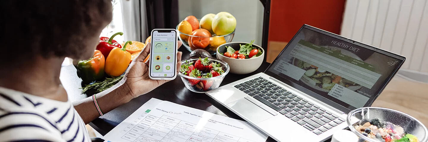 Eine Frau sitzt mit einem Smartphone am Tisch. Vor ihr liegen ein Ernährungsplan und gesunde Lebensmittel.