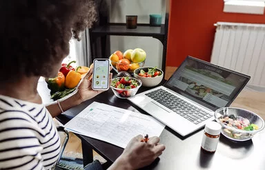 Eine Frau sitzt mit einem Smartphone am Tisch. Vor ihr liegen ein Ernährungsplan und gesunde Lebensmittel.
