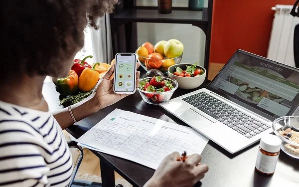 Eine Frau sitzt mit einem Smartphone am Tisch. Vor ihr liegen ein Ernährungsplan und gesunde Lebensmittel.