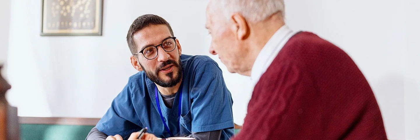 Eine männliche Pflegekraft in medizinischer Arbeitskleidung sitzt mit einem älteren Mann an einem Tisch. Die Pflegekraft lehnt sich nach vorne und blickt den älteren Mann im Profil an.