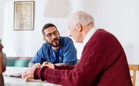 Eine männliche Pflegekraft in medizinischer Arbeitskleidung sitzt mit einem älteren Mann an einem Tisch. Die Pflegekraft lehnt sich nach vorne und blickt den älteren Mann im Profil an.