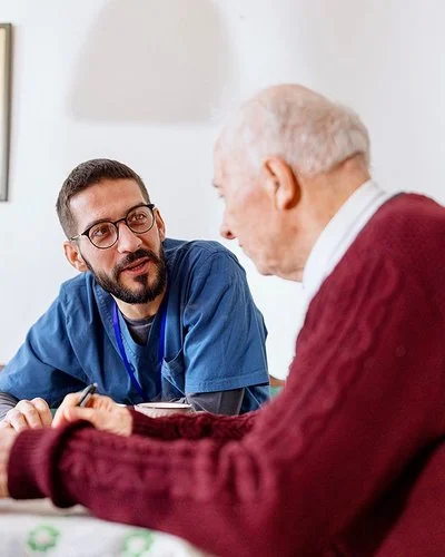 Eine männliche Pflegekraft in medizinischer Arbeitskleidung sitzt mit einem älteren Mann an einem Tisch. Die Pflegekraft lehnt sich nach vorne und blickt den älteren Mann im Profil an.