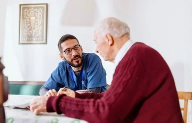 Eine männliche Pflegekraft in medizinischer Arbeitskleidung sitzt mit einem älteren Mann an einem Tisch. Die Pflegekraft lehnt sich nach vorne und blickt den älteren Mann im Profil an.