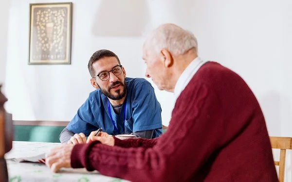 Eine männliche Pflegekraft in medizinischer Arbeitskleidung sitzt mit einem älteren Mann an einem Tisch. Die Pflegekraft lehnt sich nach vorne und blickt den älteren Mann im Profil an.