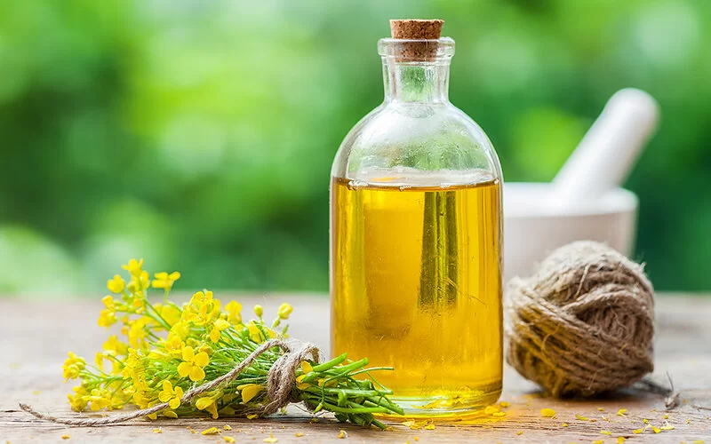 Eine Flasche Rapsöl steht auf einem Tisch, daneben liegt ein kleiner Strauß Rapsblumen, im Hintergrund ist ein Mörser zu sehen.