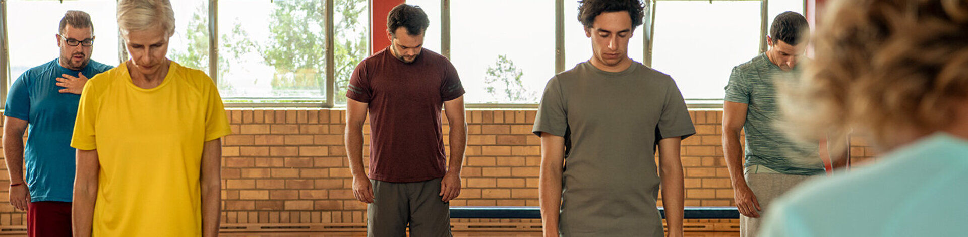Eine Frau und vier Männer stehen gegenüber einer Trainerin auf einer Yoga-Matte und halten ihren Kopf gesenkt. Die AOK unterstützt die Teilnahme an Yoga-Kursen.