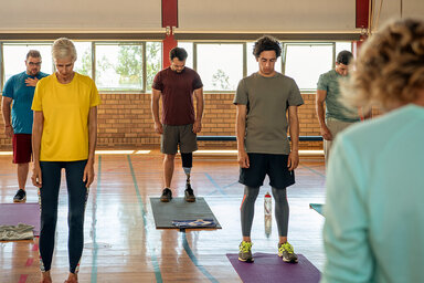 Eine Frau und vier Männer stehen gegenüber einer Trainerin auf einer Yoga-Matte und halten ihren Kopf gesenkt. Die AOK unterstützt die Teilnahme an Yoga-Kursen.