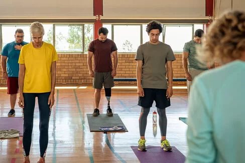Eine Frau und vier Männer stehen gegenüber einer Trainerin auf einer Yoga-Matte und halten ihren Kopf gesenkt. Die AOK unterstützt die Teilnahme an Yoga-Kursen.