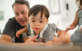 Ein kleines Kind mit Down-Syndrom wird von seinem Vater im Arm gehalten, während sie gemeinsam vor einem Laptop sitzen.