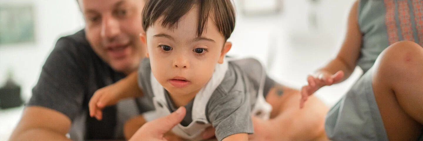 Ein kleines Kind mit Down-Syndrom wird von seinem Vater im Arm gehalten, während sie gemeinsam vor einem Laptop sitzen.