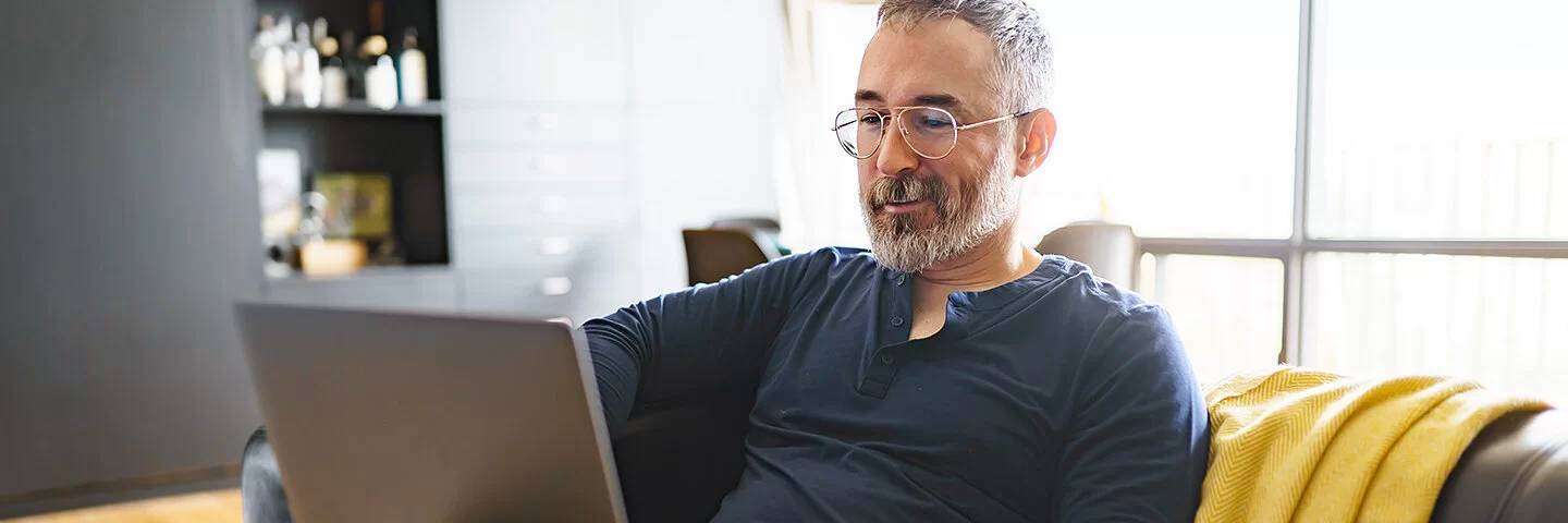 Ein Mann mittleren Alters mit kurzen grauen Haaren und Vollbart sitzt auf einem dunklen Sofa. Er sucht etwas im Internet mit einem Laptop auf seinem Schoß.