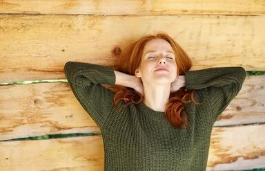 Eine junge Frau mit roten Haaren und dunkelgrünem Pullover lehnt an einer hellen Holzwand. Sie hat die Arme hinter ihrem Kopf verschränkt und die Augen geschlossen, dabei lächelt sie entspannt und zufrieden.