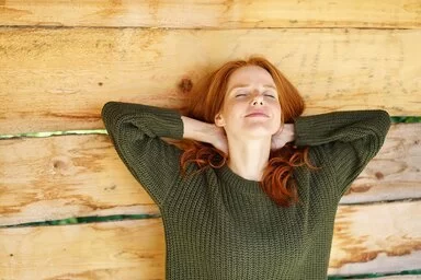Eine junge Frau mit roten Haaren und dunkelgrünem Pullover lehnt an einer hellen Holzwand. Sie hat die Arme hinter ihrem Kopf verschränkt und die Augen geschlossen, dabei lächelt sie entspannt und zufrieden.