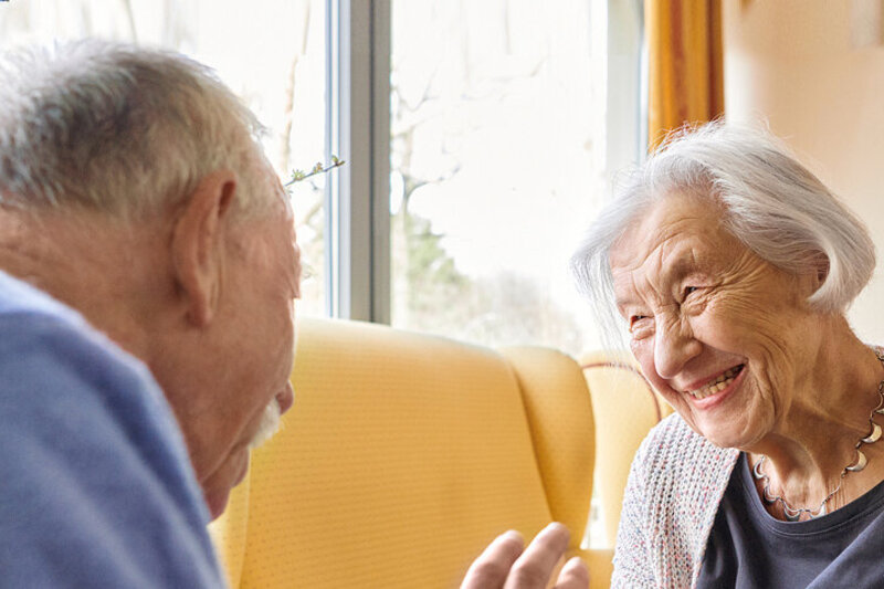 Eine ältere Frau sitzt im Wohnzimmer und lächelt ihrem Mann entgegen.