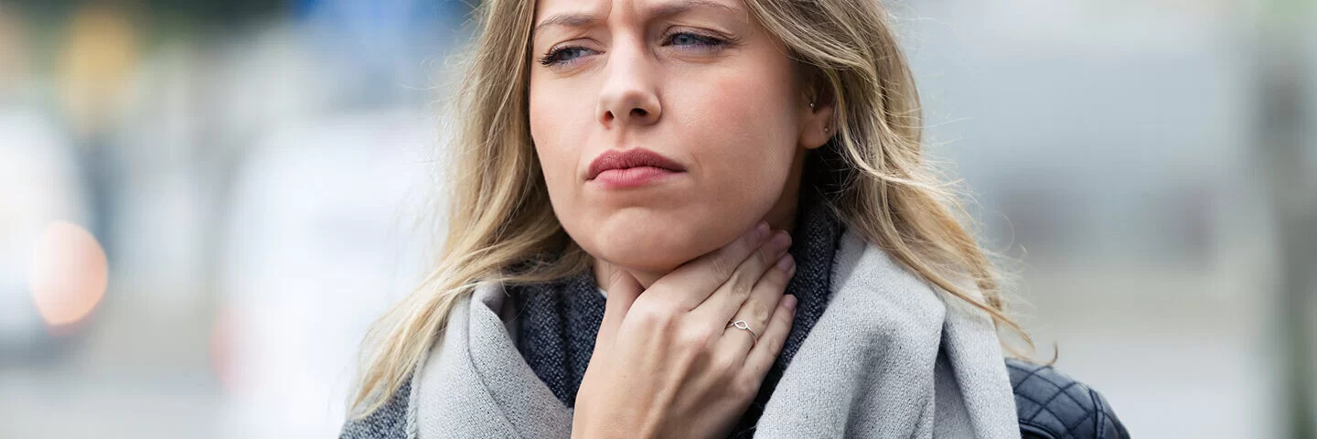 Blonde Frau mit schmerzverzerrtem Gesicht und einem Wollschal greift sich mit der rechten Hand an ihren Hals.