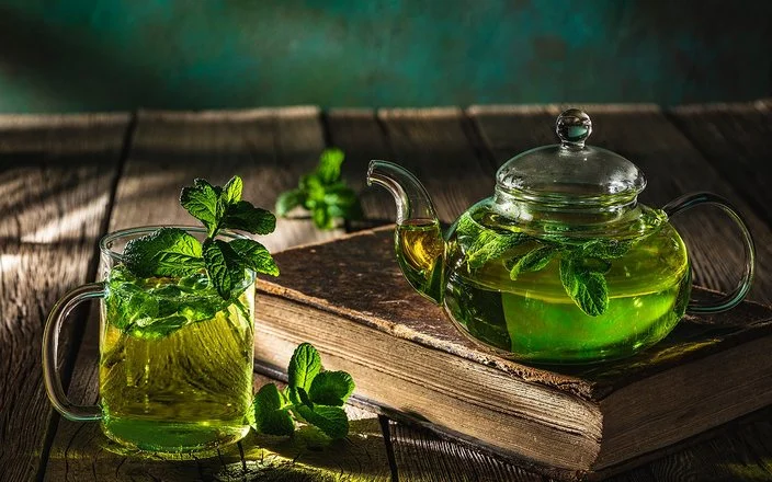 Auf einem dunklen Holztisch stehen eine Glaskanne und ein Teeglas, die beide mit frischem Pfefferminztee gefüllt sind.