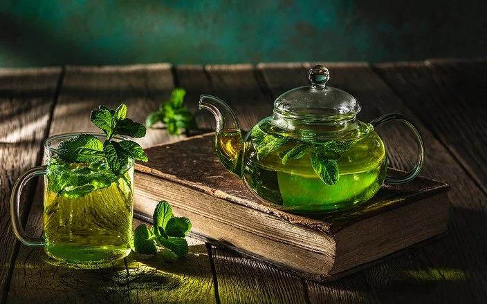 Auf einem dunklen Holztisch stehen eine Glaskanne und ein Teeglas, die beide mit frischem Pfefferminztee gefüllt sind.