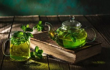 Auf einem dunklen Holztisch stehen eine Glaskanne und ein Teeglas, die beide mit frischem Pfefferminztee gefüllt sind.