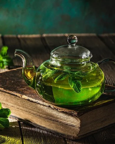 Auf einem dunklen Holztisch stehen eine Glaskanne und ein Teeglas, die beide mit frischem Pfefferminztee gefüllt sind.