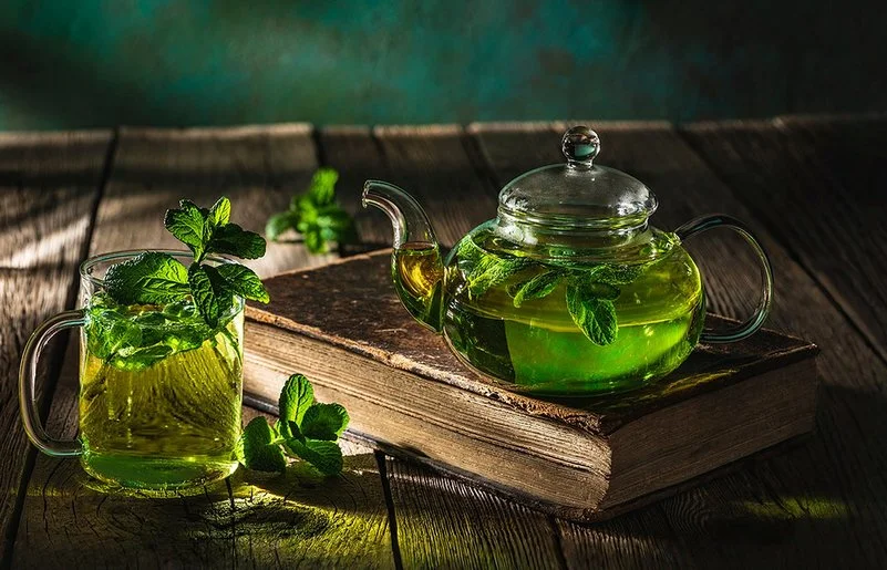 Auf einem dunklen Holztisch stehen eine Glaskanne und ein Teeglas, die beide mit frischem Pfefferminztee gefüllt sind.
