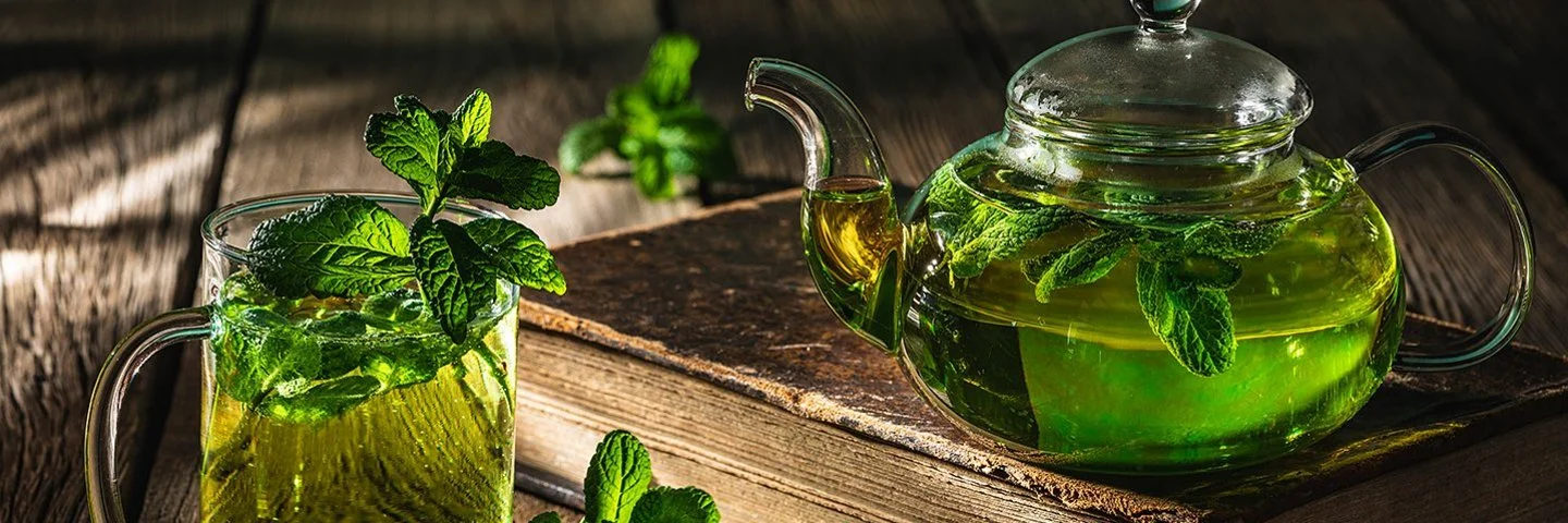 Auf einem dunklen Holztisch stehen eine Glaskanne und ein Teeglas, die beide mit frischem Pfefferminztee gefüllt sind.