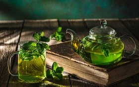 Auf einem dunklen Holztisch stehen eine Glaskanne und ein Teeglas, die beide mit frischem Pfefferminztee gefüllt sind.