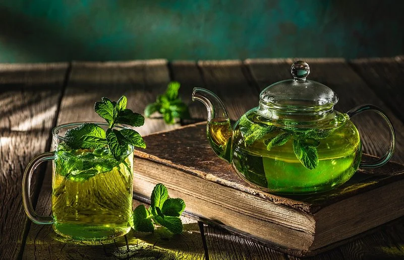 Auf einem dunklen Holztisch stehen eine Glaskanne und ein Teeglas, die beide mit frischem Pfefferminztee gefüllt sind.