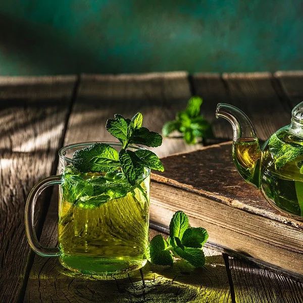 Auf einem dunklen Holztisch stehen eine Glaskanne und ein Teeglas, die beide mit frischem Pfefferminztee gefüllt sind.