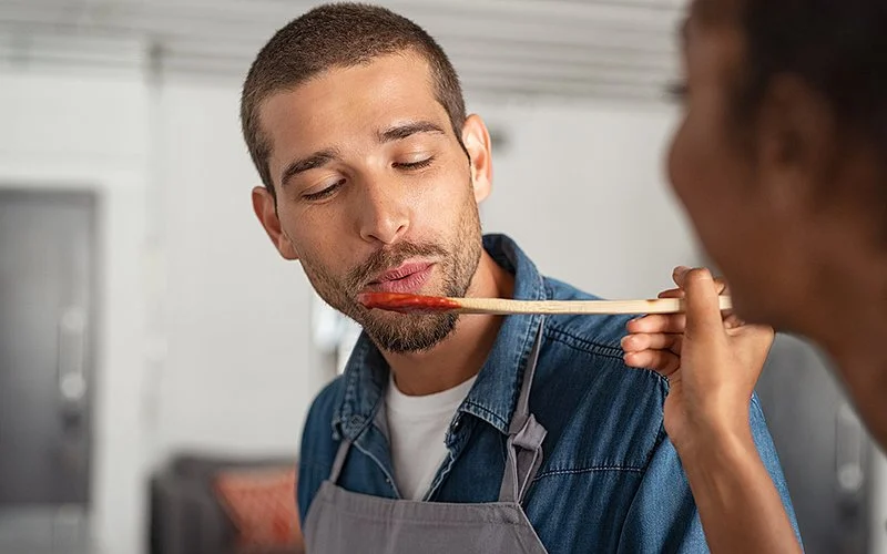 Eine junger Mann mit Schürze probiert beim Kochen Tomatensauce von einem Holzlöffel, der ihm von einer Frau neben ihm gereicht wird.