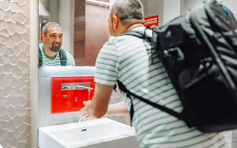 Ein mittelalter Mann steht an einem öffentlichen Waschbecken, wäscht sich die Hände und lächelt in den Spiegel.