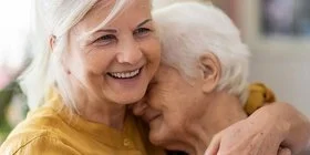 Eine jüngere Frau hält eine ältere Frau fest im Arm. Die jüngere Frau lächelt liebevoll, während sich die ältere Frau an die Schulter der anderen Frau anlehnt.