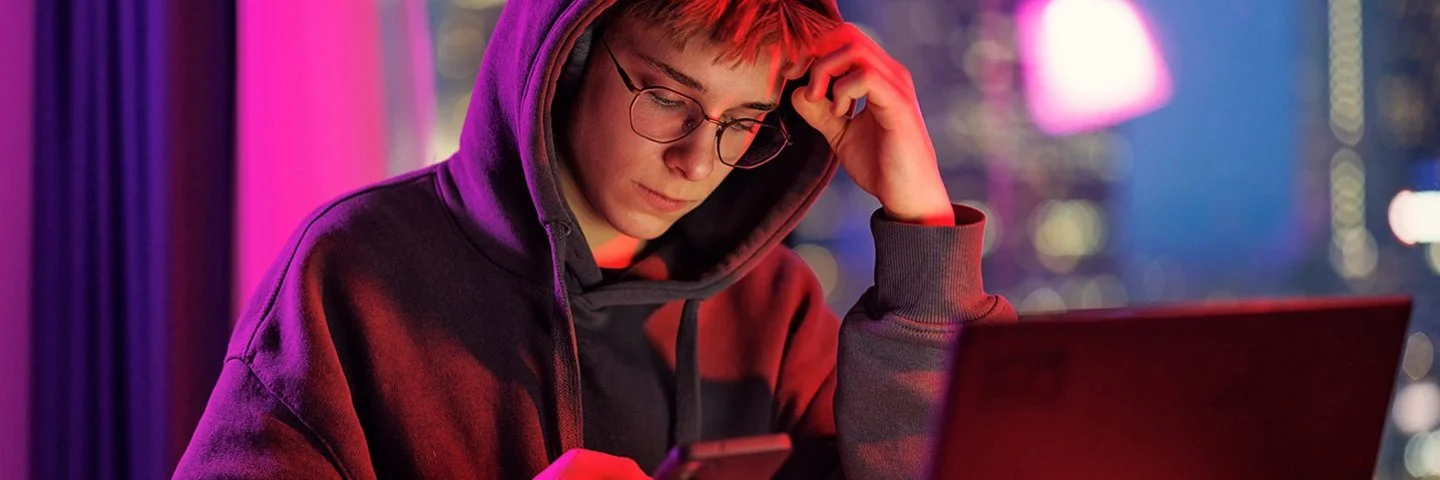 Ein mänlicher Jugendlicher in schwarzem Kapuzenpullover sitzt im Dämmerlicht vor einem Laptop, er blickt auf das Handy, das er in der Hand hält.