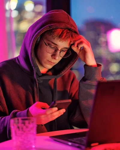 Ein mänlicher Jugendlicher in schwarzem Kapuzenpullover sitzt im Dämmerlicht vor einem Laptop, er blickt auf das Handy, das er in der Hand hält.