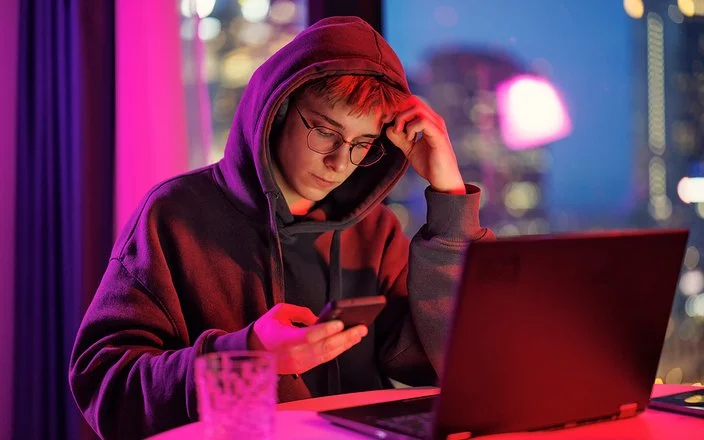 Ein mänlicher Jugendlicher in schwarzem Kapuzenpullover sitzt im Dämmerlicht vor einem Laptop, er blickt auf das Handy, das er in der Hand hält.