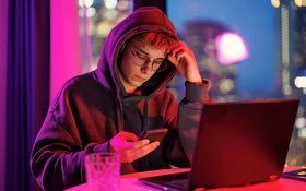 Ein mänlicher Jugendlicher in schwarzem Kapuzenpullover sitzt im Dämmerlicht vor einem Laptop, er blickt auf das Handy, das er in der Hand hält.