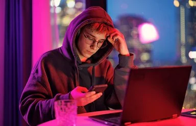 Ein mänlicher Jugendlicher in schwarzem Kapuzenpullover sitzt im Dämmerlicht vor einem Laptop, er blickt auf das Handy, das er in der Hand hält.