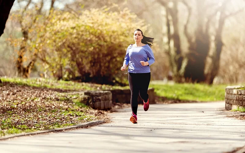 Leicht übergewichtige Frau joggt durch einen Park.