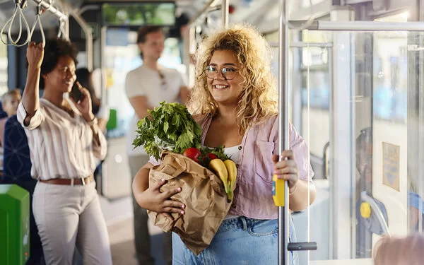 Eine Frau steht mit einer Papiertüte voll mit frischem Obst und Gemüse im Bus und hält sich an der Haltestange fest.