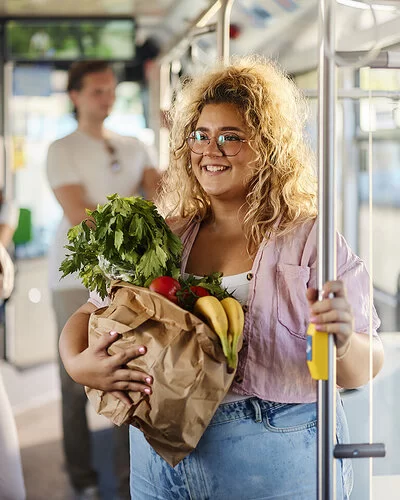 Eine Frau steht mit einer Papiertüte voll mit frischem Obst und Gemüse im Bus und hält sich an der Haltestange fest.