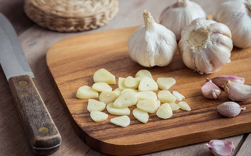 Verursacht die Symptome der Knoblauch-Unverträglichkeit: Eine geschnittene Knoblauchzehe liegt auf einem Holzbrett neben Knoblauchknollen.