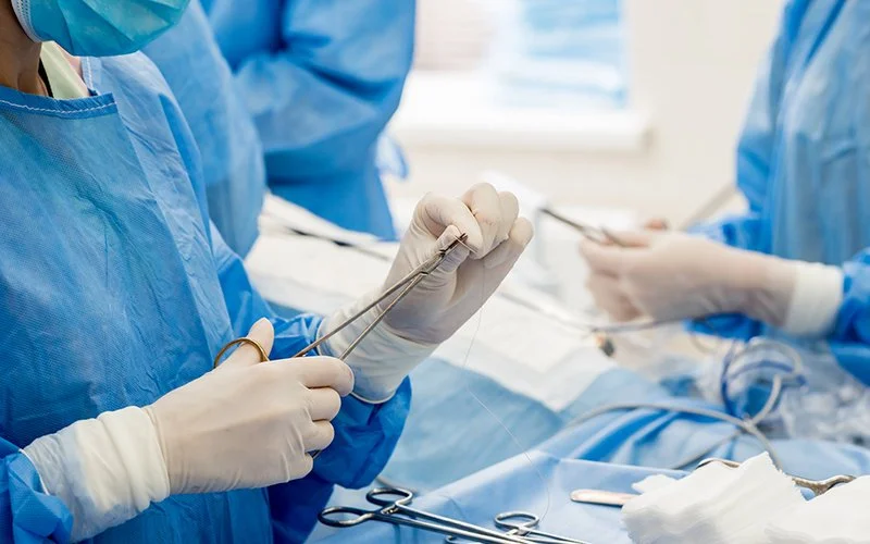 Eine Person in Schutzkleidung steht neben dem OP-Tisch und hält eine Schere und einen Faden in Händen.