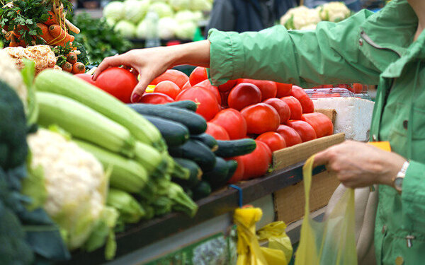 Eine Seniorin, die auf gesunde Ernährung achtet, steht auf dem Wochenmarkt vor einem Tresen mit Gemüse und greift nach einer großen Tomate.