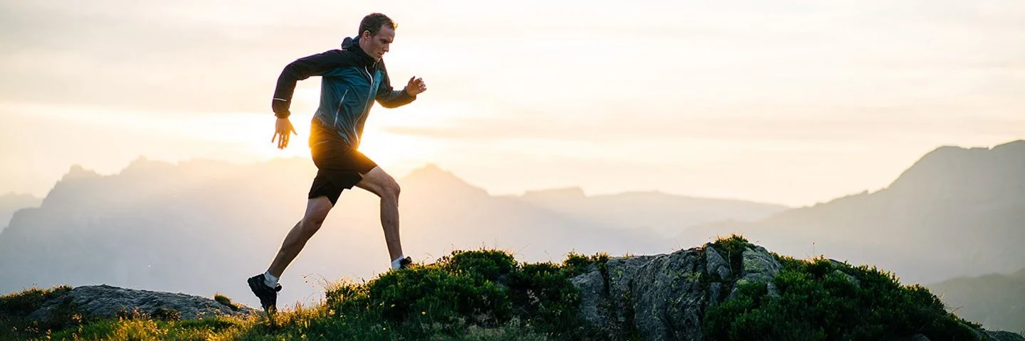Ein Mann läuft bei Sonnenaufgang in einem Gebirgsgelände. Im Hintergrund ist eine Bergkette zu sehen.