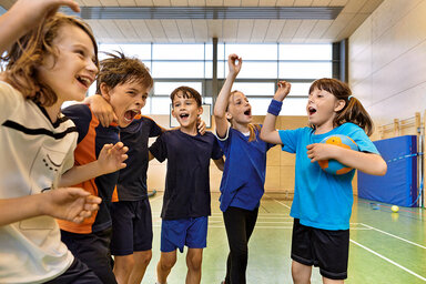 Kinder in verschiedenen Sport-Outfits fallen sich lachend in die Arme.